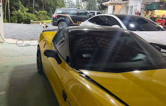 Carro esportivo amarelo em garagem, com viatura da Polícia Federal ao fundo.