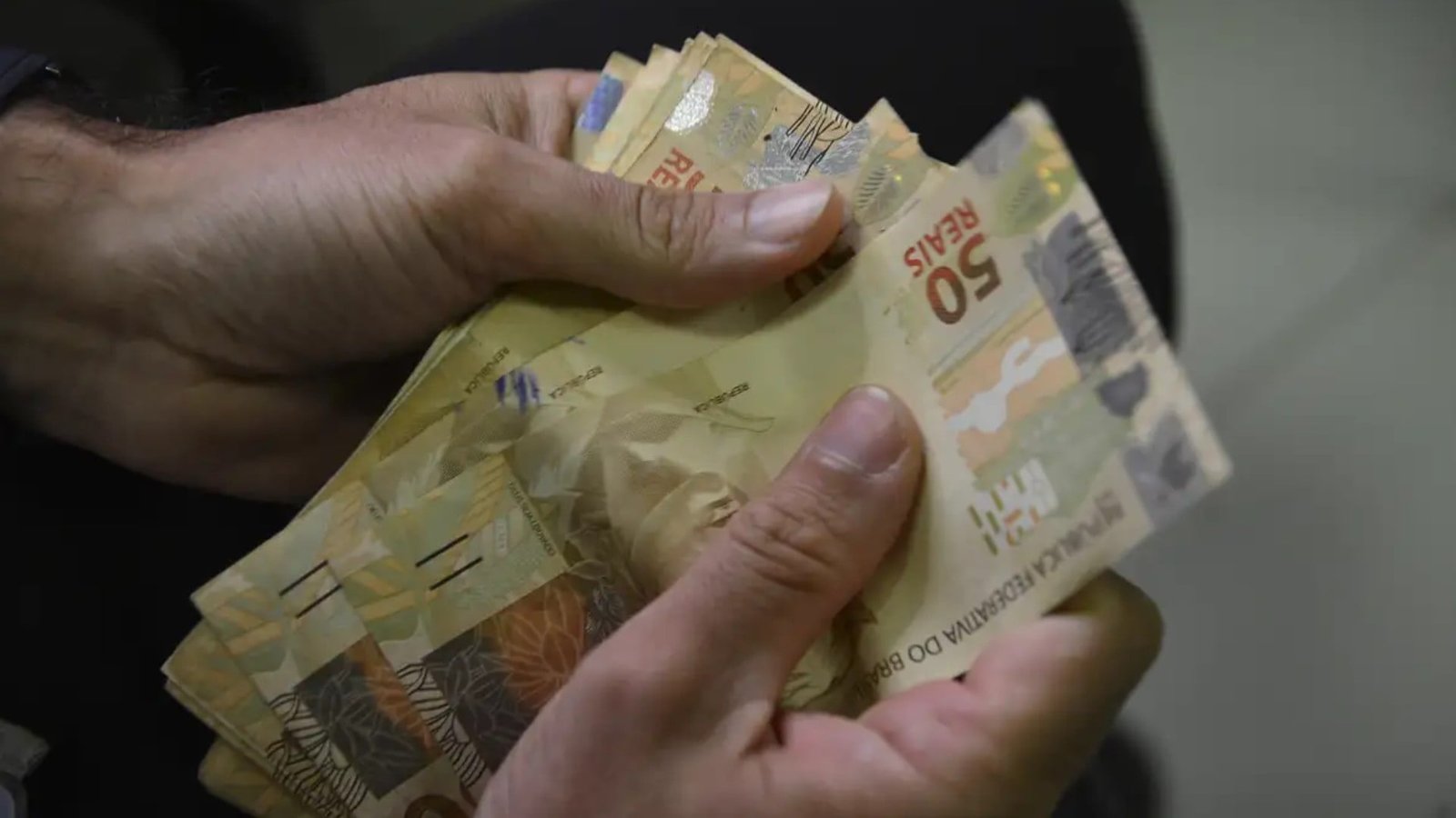 Mãos contam cédulas de cinquenta reais.
